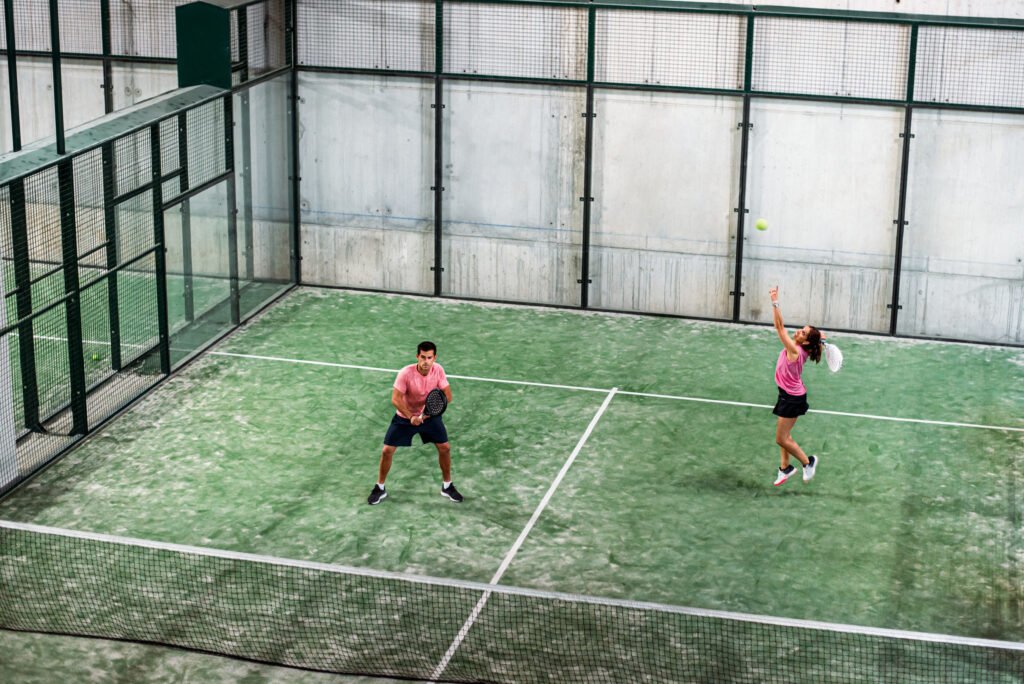 Zwei Personen spielen Padel, den beliebten Trendsport, auf einer Indoor-Anlage mit grünem Rasen. Die Frau rechts springt, um den Ball zu schlagen, während der Mann links seinen Schläger bereit macht. In rosa Oberteilen und dunklen Shorts spielen sie in einem Gehäuse aus Glas und Metall.