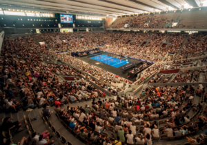 Eine große Indoor-Arena voller Zuschauer, die sich ein Padel-Match auf einem blauen Platz ansehen. Die Tribünen sind voll und die Atmosphäre ist lebhaft. Helle Lichter erhellen den Veranstaltungsort und die Anzeigetafel ist über dem Platz sichtbar.