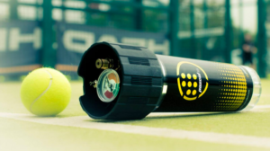 Ein Tennisball liegt auf einem Platz neben einem Druckbehälter, der dafür gemacht ist, ihn frisch zu halten. Der schwarze Behälter, verziert mit einem gelben Logo, steht fest. Im Hintergrund unterstreicht ein unscharfer Tennisplatzzaun subtil die Ruhe der Szene.
